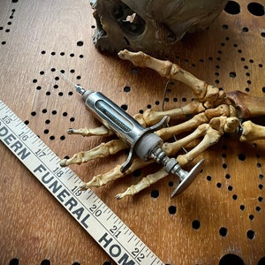 May include: An antique silver syringe rests on a skeletal hand, with a wooden ruler reading "MODERN FUNERAL HOME" in the foreground. A skull fragment is visible in the background. The items are on a wooden surface.