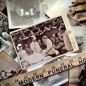 May include: A collection of vintage photographs, including a black and white photo of a group of people in a medical setting, a photo of a cemetery, and a photo of a family standing on a porch. A wooden ruler with the words "Modern Funeral Home" is visible in the foreground.