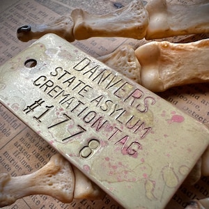 May include: A weathered brass tag with the text "DANVERS STATE ASYLUM CREMATION TAG #1778" is displayed. The tag has a distressed finish with pink accents and is set against a backdrop of aged paper and bone fragments.