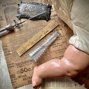 Peut inclure: Une seringue médicale ancienne, un dispositif en plastique transparent et une règle en bois reposent sur un fond de journal vintage. Un bras de poupée est également présent. L'ensemble évoque l'histoire et la nostalgie.