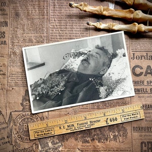 May include: A black and white photograph of a man lying in a casket, surrounded by flowers. The photo is pinned to a piece of paper with a wooden ruler and the text "R.C. Nein, Funeral Director, Front & Dayton Street, Hamilton, Ohio" printed on it.