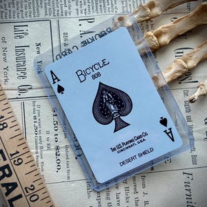 May include: A Bicycle 808 Ace of Spades playing card in a protective sleeve. The card features a black spade design with the text "Desert Shield" and "The US Playing Card Co." on a vintage newspaper background.
