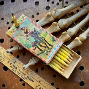 May include: A rectangular matchbox featuring a vintage illustration of a person riding a donkey, labeled Ritorno dal Mercato, containing yellow wax tapers. A wooden ruler measuring 6 inches is placed alongside, resting on a skeletal hand model.