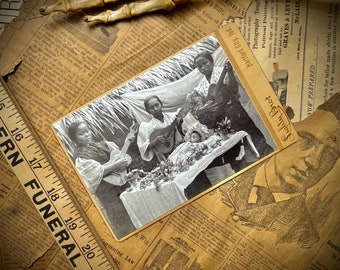 May include: A black and white photograph of four people playing musical instruments. Three women are playing guitars and a baby is lying on a table covered in flowers. The photo is framed with a gold border and has the text "Hartford City, Ind." and "Photography Tangl" printed on it.