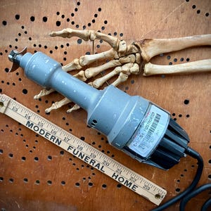 May include: A gray and black power tool with a blade, labeled "POSTMORTEM", rests on a wooden surface. A wooden ruler with the text "MODERN FUNERAL HOME" and a human skeleton hand are also present. The tool has a black power cord.