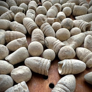 May include: A collection of antique lead bullets and round balls, in various shapes and sizes, scattered on a wooden surface. The bullets have a weathered, aged appearance, with a light gray color.