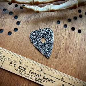 May include: A silver-colored planchette pendant with a Ouija board design. The pendant features the alphabet, numbers, and the words "YES", "NO", and "GOODBYE". The pendant is on a wooden surface with a ruler.