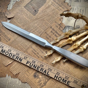 May include: A stainless steel letter opener with a pointed blade and rounded handle rests on vintage newspaper. A wooden ruler with "MODERN FUNERAL HOME" printed on it is in the foreground. A skeletal hand is in the background.