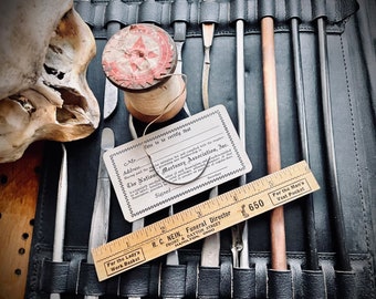 Puede incluir: Una caja de cuero negro con un juego de herramientas, que incluye una regla de madera con el texto "For the Lady's Work Basket" y "For the Man's Vest Pocket", una tarjeta blanca con el texto "The National Mortuary Association, Inc." y una bobina de hilo blanco con una estrella roja.