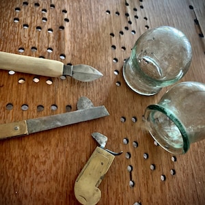 May include: An overhead shot shows antique medical tools and two glass cupping jars on a wooden surface. The tools include a scalpel with a bone handle, and two other tools with metal blades and brass or metal handles. The cupping jars are clear glass.