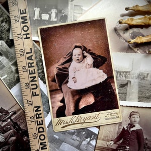May include: A collection of vintage photographs, including a portrait of a young child in a white dress, a photo of a cemetery, and a photo of a man in a suit. The photos are arranged on a wooden surface with a ruler that reads "Modern Funeral Home".