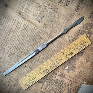 May include: A silver metal surgical scalpel with a sharp blade and a wooden ruler with the text "For the Man's Vest Pocket" and "650" printed on it.