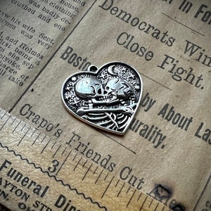 May include: A silver heart-shaped pendant with a skeletal couple embracing. The pendant features a crescent moon and stars.