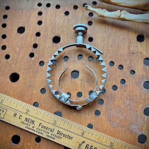Puede incluir: Un dispositivo circular de metal plateado con un borde exterior dentado y un mecanismo central de resorte, colocado junto a una regla de madera que muestra un diámetro de aproximadamente 9 centímetros para el componente circular.