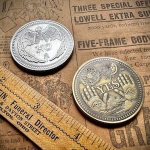 Puede incluir: Dos fichas de metal plateadas y doradas con diseños grabados. La ficha plateada tiene un esqueleto con alas y la palabra "NO" en el centro. La ficha dorada tiene dos manos con ojos y la palabra "YES" en el centro.