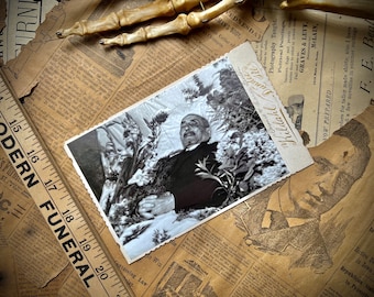 May include: A black and white photograph of a man lying in a casket surrounded by flowers. The photo is on a table with old newspapers and a skeleton hand.