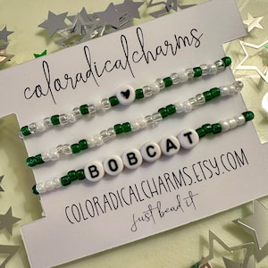 May include: Three white and green beaded bracelets with black letter beads spelling "BOBCAT" and a small black heart charm. The bracelets are displayed on a white card with the text "coloradicalcharms just bead it COLORADICALCHARMS.ETSY.COM"