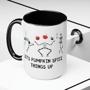 May include: White ceramic mug with a black interior and handle. The mug features a design of dancing skeletons, red pumpkins, and the text "Let's Pumpkin Spice Things Up" in black. A fun, seasonal item for Halloween.