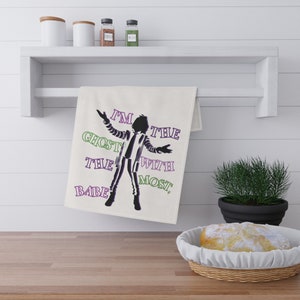 May include: A cream-colored kitchen towel with the text "I'M THE GHOST WITH THE MOST, BABE" in green and purple. The towel features a black and white silhouette of a character with arms outstretched. The towel is hanging on a white shelf.