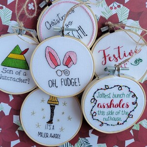 May include: A set of 6 embroidered Christmas ornaments with various holiday-themed designs. The ornaments feature phrases like "Son of a Nutcracker!" and "It's a Major Award!"
