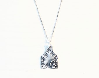 May include: A silver necklace with a pendant shaped like a livestock tag. The tag has a geometric pattern of diamonds and a small flower design.