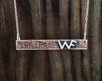 Puede incluir: Un collar de cadena de plata con un colgante de barra de cobre. La barra tiene un diseño floral y las iniciales "WF" en plata.