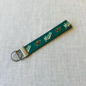 May include: A green and white Minnesota Wild keychain wristlet. The wristlet features the team logo and the word "Minnesota" in white. The keychain has a silver metal ring and a silver metal clasp.