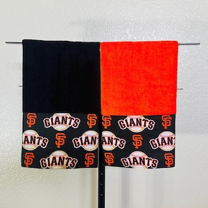 May include: Two towels, one black and one orange, are displayed. Each towel has a decorative band featuring baseballs and the text "GIANTS" in white on a black background. The towels are hanging on a black metal bar.