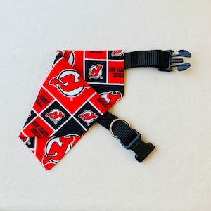 May include: Red and black dog collar featuring a New Jersey Devils logo and a black buckle. The collar is made of fabric and has a black strap.