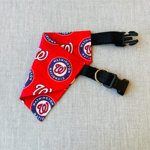 Puede incluir: Bandana roja para perros con el logotipo de los Nacionales de Washington en blanco y azul. La bandana tiene un collar negro ajustable con una hebilla de plástico y un anillo de metal dorado.