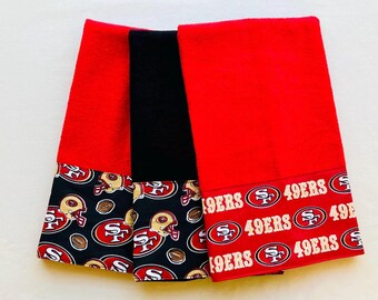 Op de afbeelding: Set van drie keukendoeken in rood en zwart met een San Francisco 49ers-thema. De handdoeken hebben een zwart-witte print van voetbalhelmen en voetballen met de woorden "49ers" en "SF" aan de onderkant.