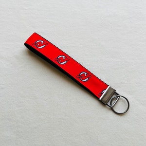 May include: Red and black key chain with a repeating pattern of a white hockey team logo. The key chain has a silver metal ring.