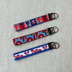 May include: Three fabric keychains featuring Philadelphia Phillies baseball team branding. The top strap has a red, white, and blue chevron pattern with team logos. The middle strap is blue with red circular logos. The bottom strap is white with red text reading Phillies.