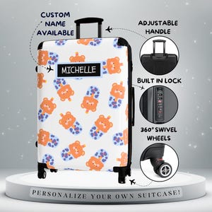 May include: White suitcase with a repeating pattern of orange teddy bears and blue polka dots. Features include an adjustable handle, built-in lock, and 360° swivel wheels. The name "MICHELLE" is displayed on a black label. The text "CUSTOM NAME AVAILABLE" is also visible.