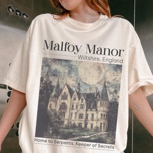 May include: Ivory coloured t-shirt featuring a graphic print of Malfoy Manor, a large, dark, gothic-style house with a full moon in the background. The text "Malfoy Manor, Wiltshire, England" and "Home to Serpents, Keeper of Secrets" is printed on the shirt.