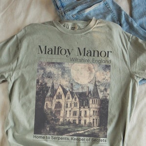 May include: A green t-shirt featuring a graphic of a large, grey stone manor house with a full moon in the sky. The text "Malfoy Manor" is above the image and the text "Wiltshire, England" is below the image. The text "Home to Serpents, Keeper of Secrets" is below the manor house.