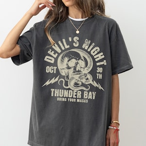 May include: A dark gray t-shirt with a graphic print of a skull with a snake wrapped around it. The text "Devil's Night Oct 30th Thunder Bay Bring Your Masks" is printed on the shirt.