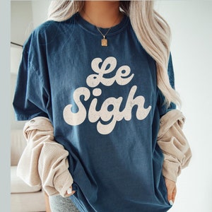 May include: A dark blue oversized t-shirt with the text "Le Sigh" in a white retro font.
