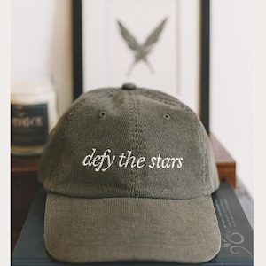 Könnte beinhalten: Olivgrüne Cord-Baseballkappe mit dem weißen Schriftzug "defy the stars". Die Kappe hat einen gebogenen Schirm und eine runde Krone. Die Mütze liegt auf einem Bücherstapel.