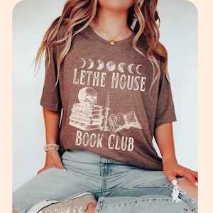 May include: A brown t-shirt with a white graphic design featuring a skull, books, and a candle. The text on the shirt reads "Lethe House Book Club".
