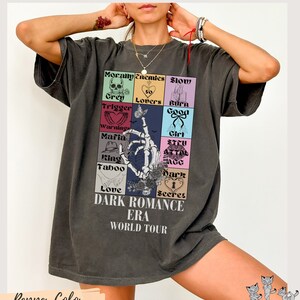 May include: A dark gray t-shirt with a graphic print of a skeleton hand holding a bouquet of flowers. The graphic includes the text "Dark Romance Era World Tour" and a grid of squares with text and images. The squares include the text "Morally Grey", "Enemies", "Slow Burn", "Trigger Warning", "Lovers", "Good Girl", "Mafia King", "Stay Alive", "Taboo Love", "Dark Secret".
