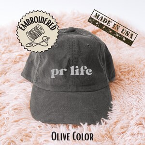 Op de afbeelding: Olijfgroene corduroy baseballcap met wit geborduurd "pr life". De cap heeft een gebogen klep en een gestructureerde kroon. De tekst "Embroidered" en "Made in USA" is ook zichtbaar.