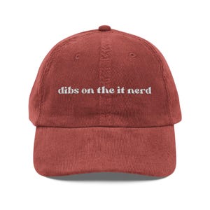 May include: A rust-colored corduroy baseball cap featuring the embroidered white text "dibs on the it nerd" centered on the front panel. The cap has a structured crown, a curved brim, and a button at the top, showcasing a textured fabric finish.