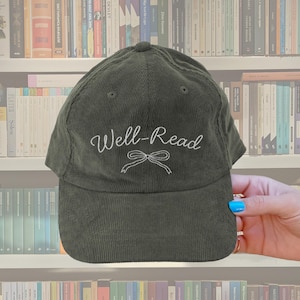 May include: An olive green corduroy baseball cap featuring the embroidered text "Well-Read" in white script above a bow. The cap has a curved peak and a rounded crown. The background shows a bookshelf filled with books.