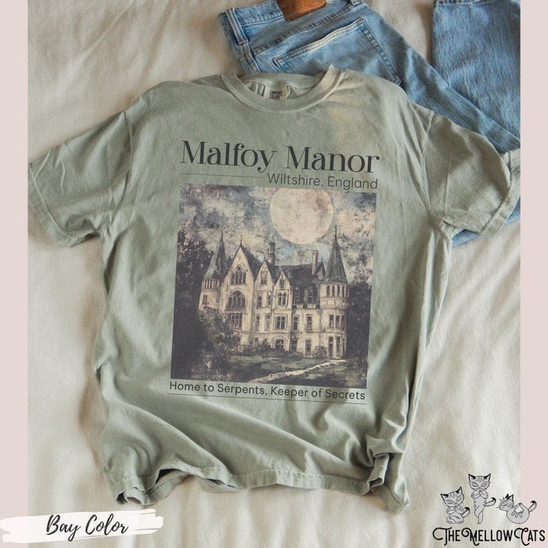 May include: A green t-shirt featuring a graphic of a large, grey stone manor house with a full moon in the sky. The text "Malfoy Manor" is above the image and the text "Wiltshire, England" is below the image. The text "Home to Serpents, Keeper of Secrets" is below the manor house.