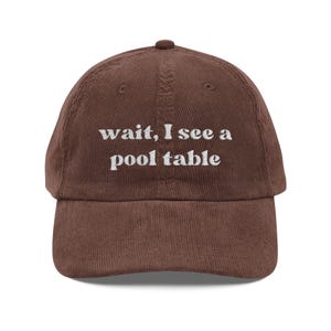 May include: A brown corduroy baseball cap with the embroidered text "wait, I see a pool table" in white. The cap has a curved brim and a button on top. This hat is a great accessory for casual wear.