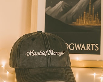 May include: A dark gray baseball cap with the embroidered text "Mischief Managed" in white script. The cap has a curved brim and a button on top. The background includes a framed poster with a mountain and castle scene.