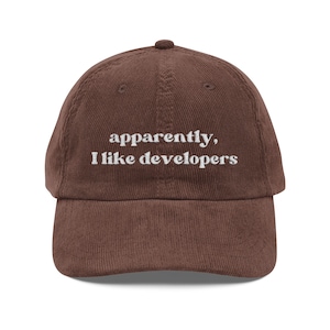 May include: A brown corduroy baseball cap with a curved brim. The cap features the embroidered text "apparently, I like developers" in white, centered on the front. The cap has a rounded top and an adjustable strap.