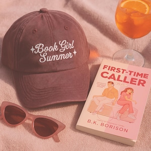 May include: A mauve baseball cap with the text "Book Girl Summer" embroidered in white, next to a paperback book titled "First-Time Caller" and pink sunglasses. A glass of orange cocktail is also in the image.