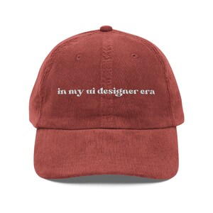 Pode incluir: Boné de beisebol de veludo cotelê vermelho tijolo com aba curva. O boné apresenta o texto bordado "in my ui designer era" em branco. O chapéu tem um design clássico de seis painéis e uma alça ajustável.
